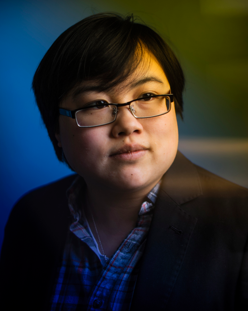 Headshot of Lydia Brown, young East Asian person, with stylized blue and yellow dramatic background. They are looking in the distance and wearing a plaid shirt and black jacket. Photo by Adam Glanzman.