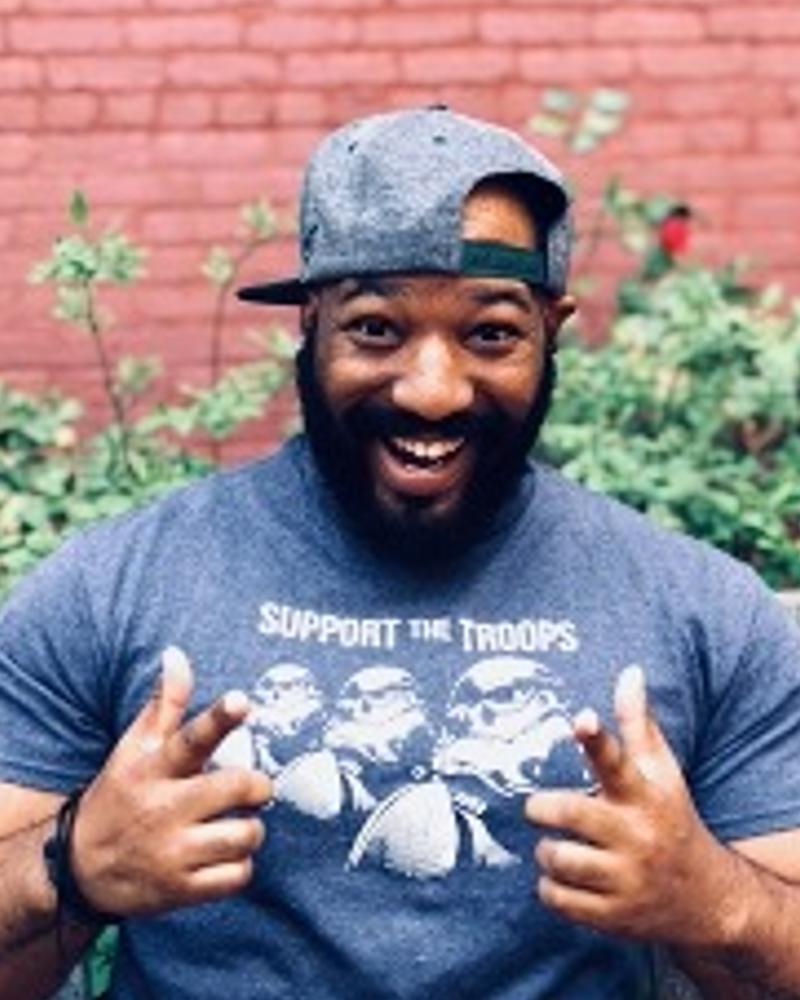 Picture of a smiling, Black, bearded male with fingers pointing at the camera in front of a brick wall with foliage. He is wearing a charcoal hat backwards with a charcoal shirt that says 
