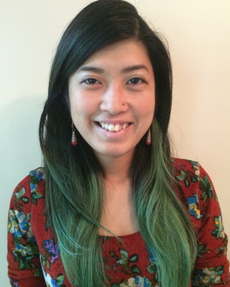 North East Asian feminine person with a nose pierce is smiling. Her long hair is half turquoise in the bottom and half black in the top. She is wearing a red sweater and red and silver earrings.