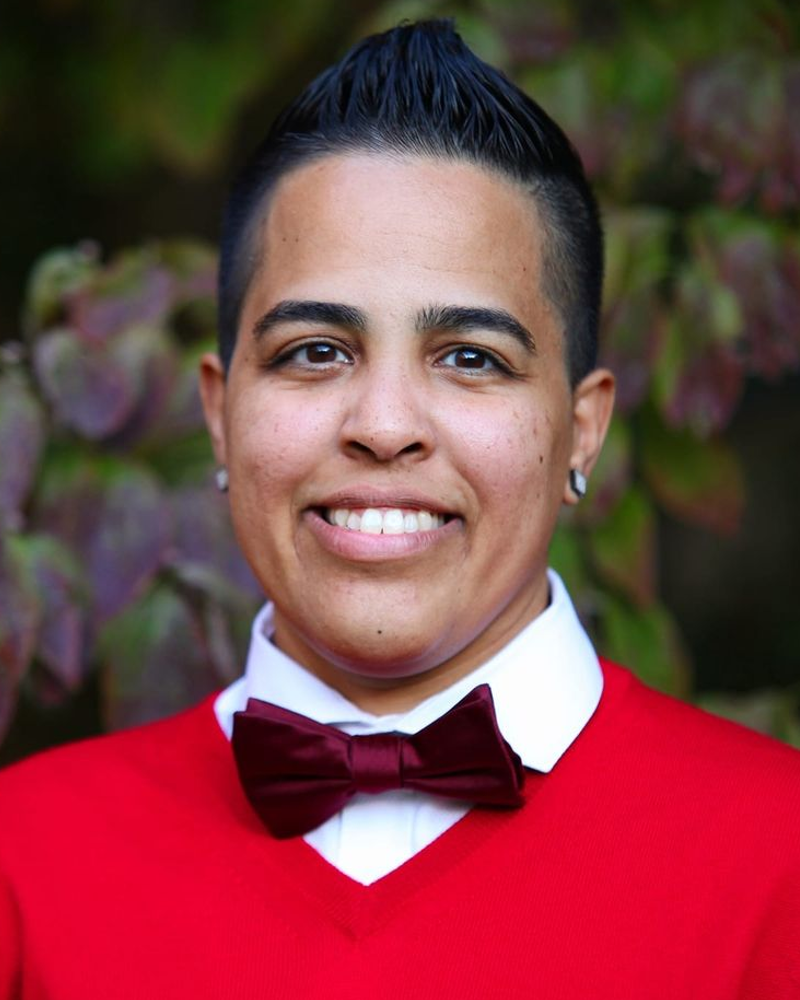 A brown, latinx, non-binary person with short black hair, wearing a red sweater, white dressy shirt underneath, stud earrings, and a black bow tie. They are smiling and behind them are faded green plants. 