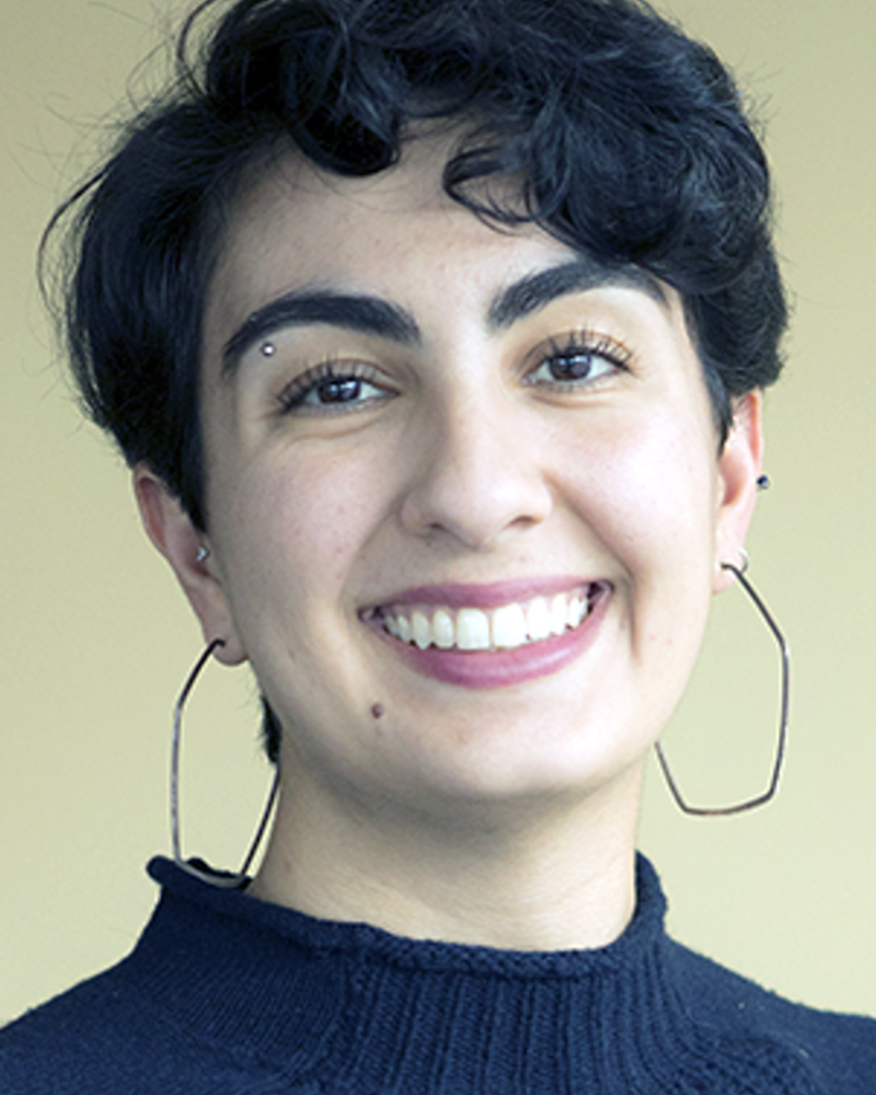 A woman with olive skin and short black curly hair smiles at the camera wearing a navy blue sweatshirt, large hoop hearings, and an eyebrow piercing.