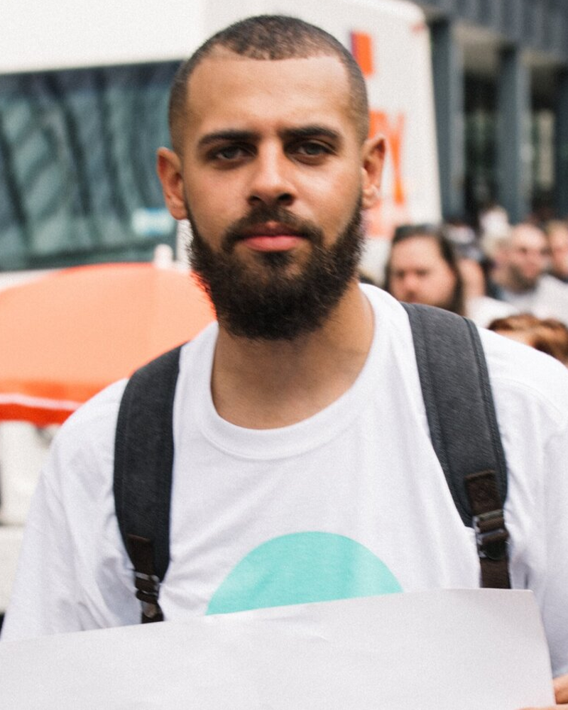 Dustin, a light skin Black man, stands in the middle of the photo looking directly into the camera. He has dark hair, a low cut fade with a part and a dark beard. He is wearing a backpack and a white tee-shirt that has an aqua blue circular shape on it. Behind him is a blurry crowd of people, a delivery truck, a building and tree as well as a traffic sign. The people in the crowd are all facing the same direction and at least one of them is holding a sign.