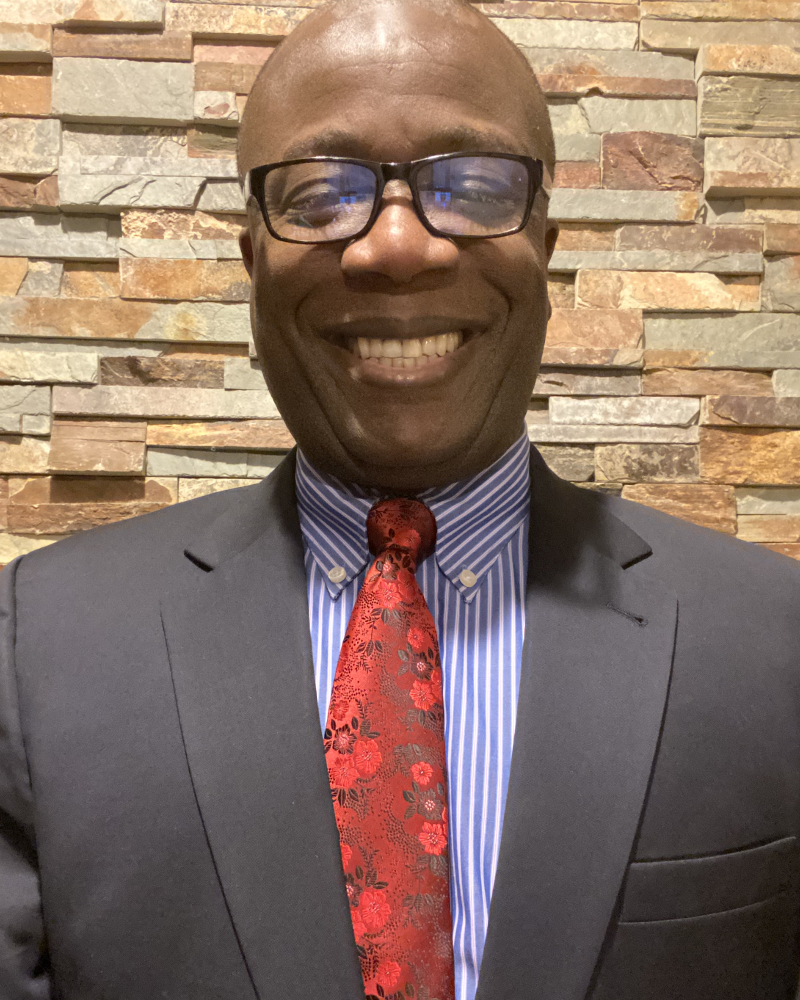 Kojo is dressed up in a navy blue suit over a blue stripped shirt and a red tie wearing reading glasses and smiling.