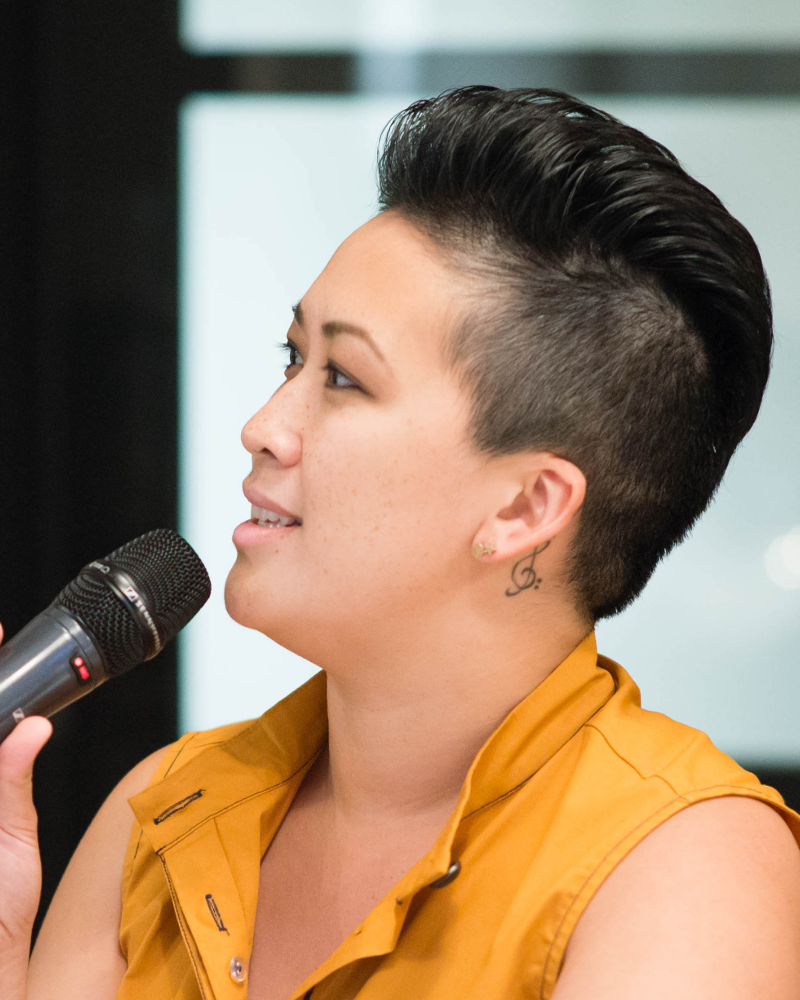 an Asian American, Vietnamese American, queer femme-ish woman in her late 30s with short black hair and light tan/olive skin looking towards the left of the photo and speaking into a handheld microphone, a small tattoo of a combined treble and bass clef behind her ear.