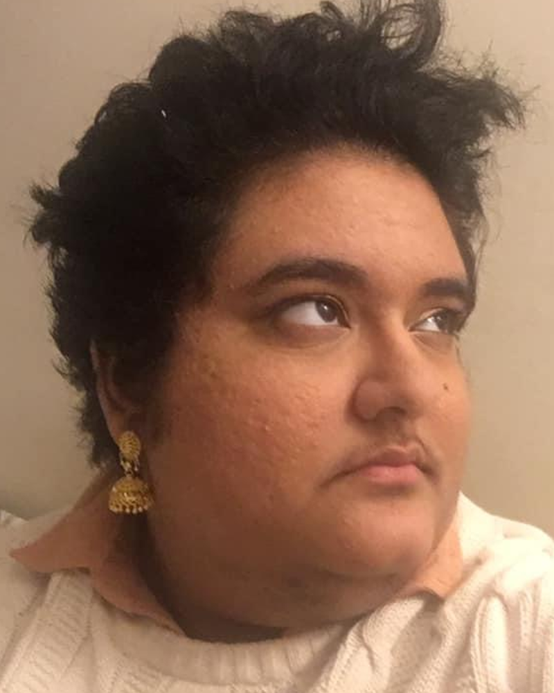 A south Asian non-binary person looks up and to the right. He wears a white sweater with a dusty pink collar peeking out, minimal makeup, and gold jhumka earrings