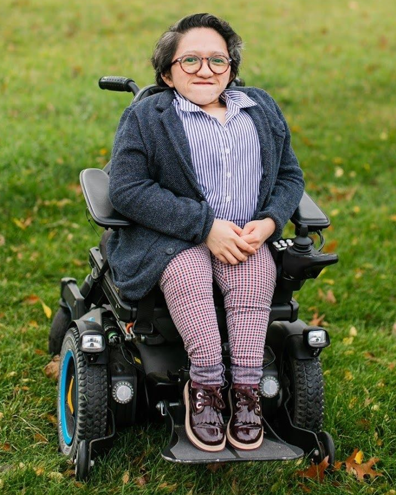 This is a photo of Sandy Ho who is a short statured queer Asian American woman sitting in a power wheelchair. She is wearing a gray unbuttoned sweater, a blue and white striped shirt, maroon shoes, and red checkered pants. She has short dark hair and glasses. Around her is a field of green grass and there are houses and trees in the background.