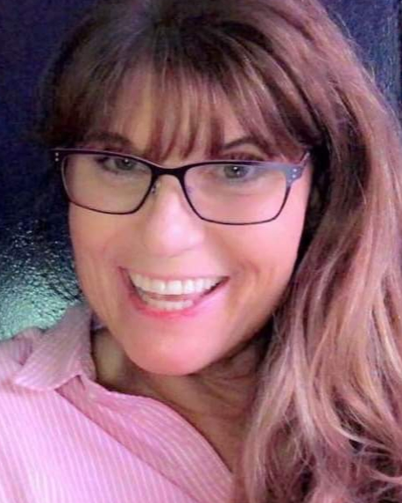 a headshot showing Sharon wearing large square glasses and long brown hair, looking directly at the camera and smiling widely and warmly. They are wearing a light pink collared shirt.