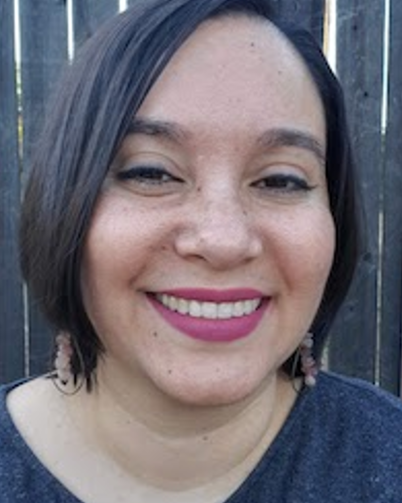 Dr. Shayda Kafai, a light, brown-skinned femme with straight hair that ends above her shoulders smiles widely into the camera. She is wearing bright magenta lipstick and rose quartz earrings. She is wearing a plain, dark grey shirt and is sitting in front of a wooden fence.