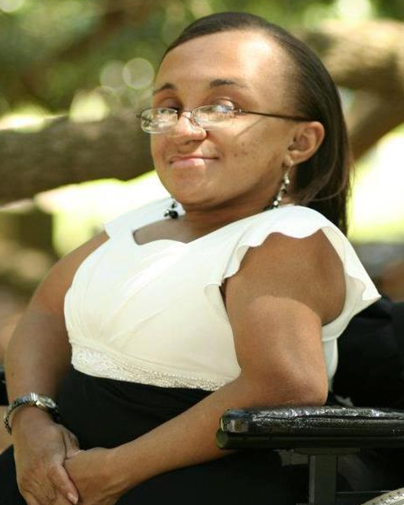 Photo of a young Black woman with her hair parted and hanging down straight. She is smiling and facing the camera sideways in her wheelchair. She is outside under the tree branches while wearing a white and black dress.