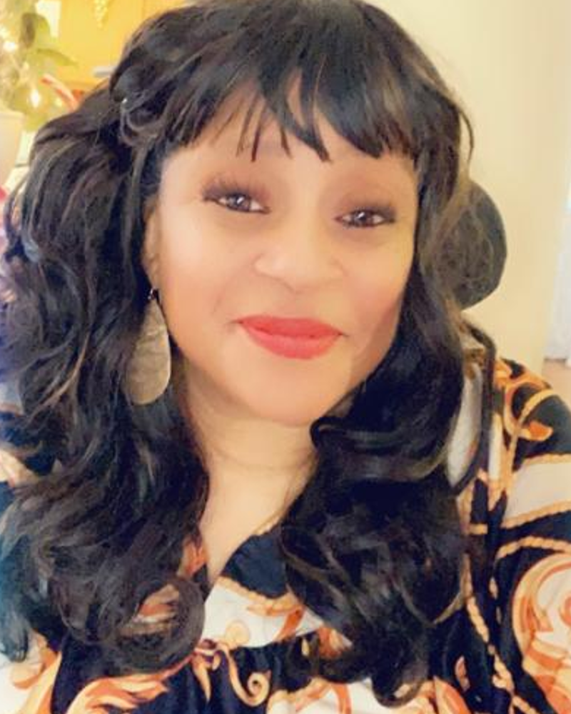 I am an African American woman with long wavy reddish-brown hair in a bob style. I have a smile on my face and wearing red lipstick. I am wearing a black, white, and gold pattern dress. I am also wearing long gold earrings.