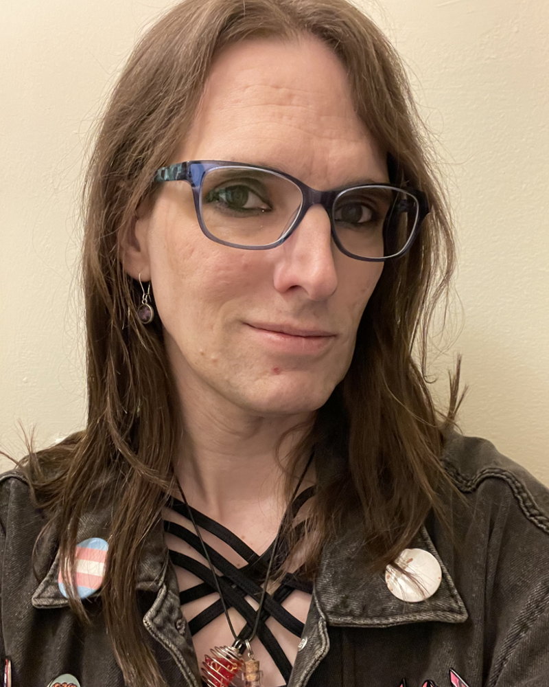 A young woman with glasses looking at the camera, she has long hair and is wearing a black jean jacket with some buttons and pins visible, one of which is the trans flag.