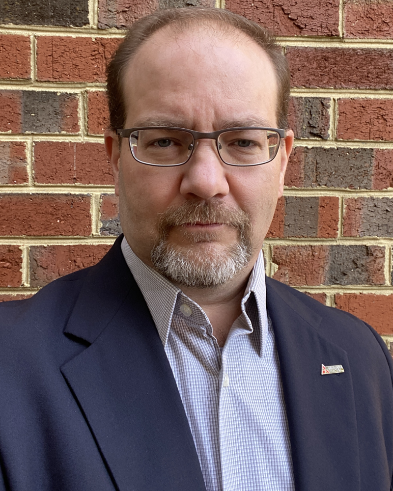 A white male with a beard wearing a blue jacket and glasses standing in front of a brick wall.