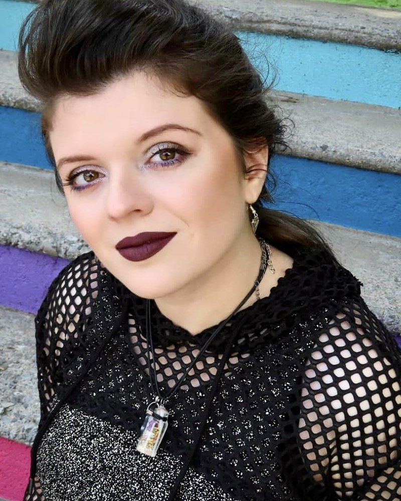 Marie, a pale skinned woman with her dark hair pulled back into a fashionable braid, sits on a set of rainbow stairs looking up towards the camera. She is wearing a cropped black fishnet hoodie over a black and silver dress paired with a dark lip.