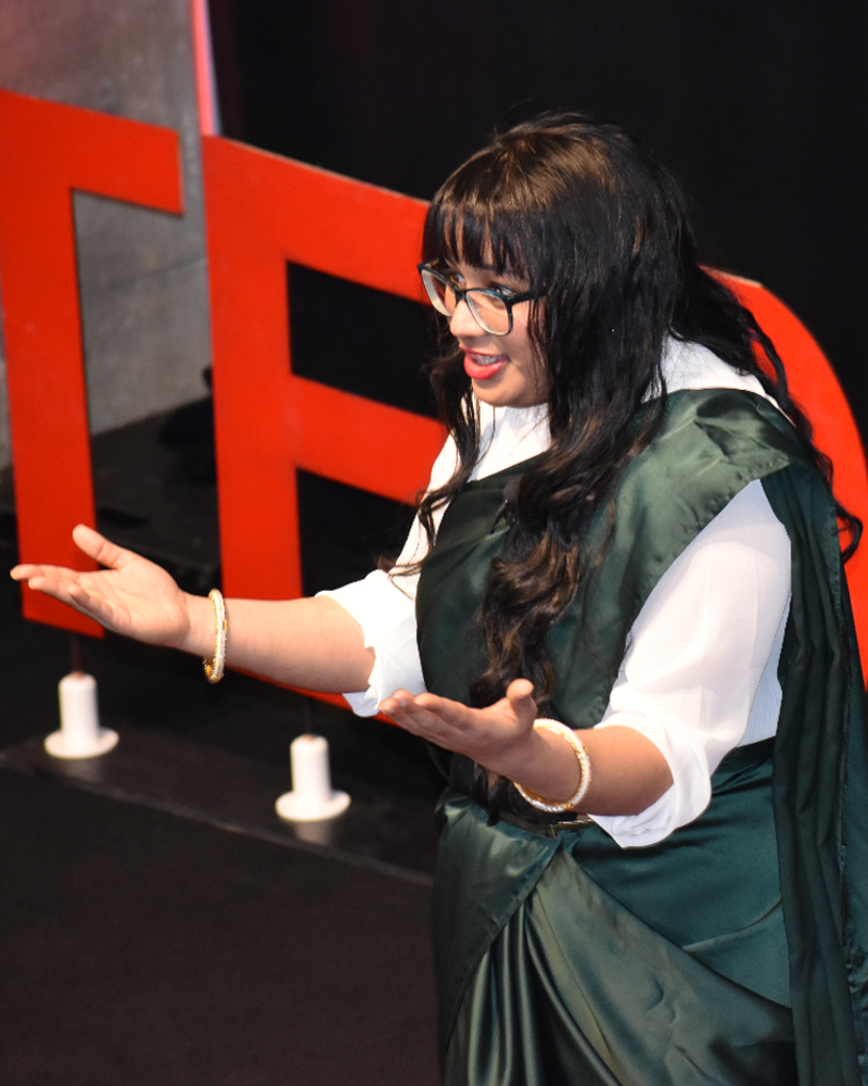 Nandita Gupta wearing a emerald, green sari with her hands outstretched, on a stage with the words 'TEDx Georgia Tech' in the background.