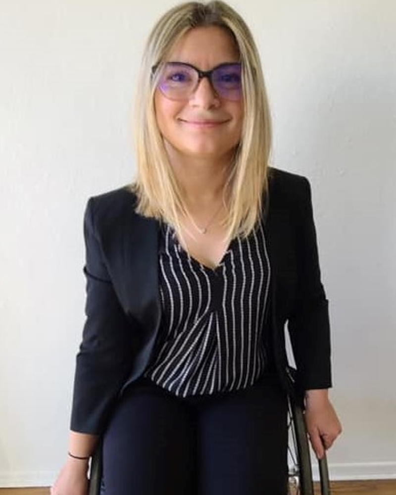 White woman, wheelchair user smiling (posed) in front of a white wall, pink/clear framed glasses with short blonde hair wearing a black blazer with a black and white stripped shirt/black pants hands are placed on the rim of the wheelchair.