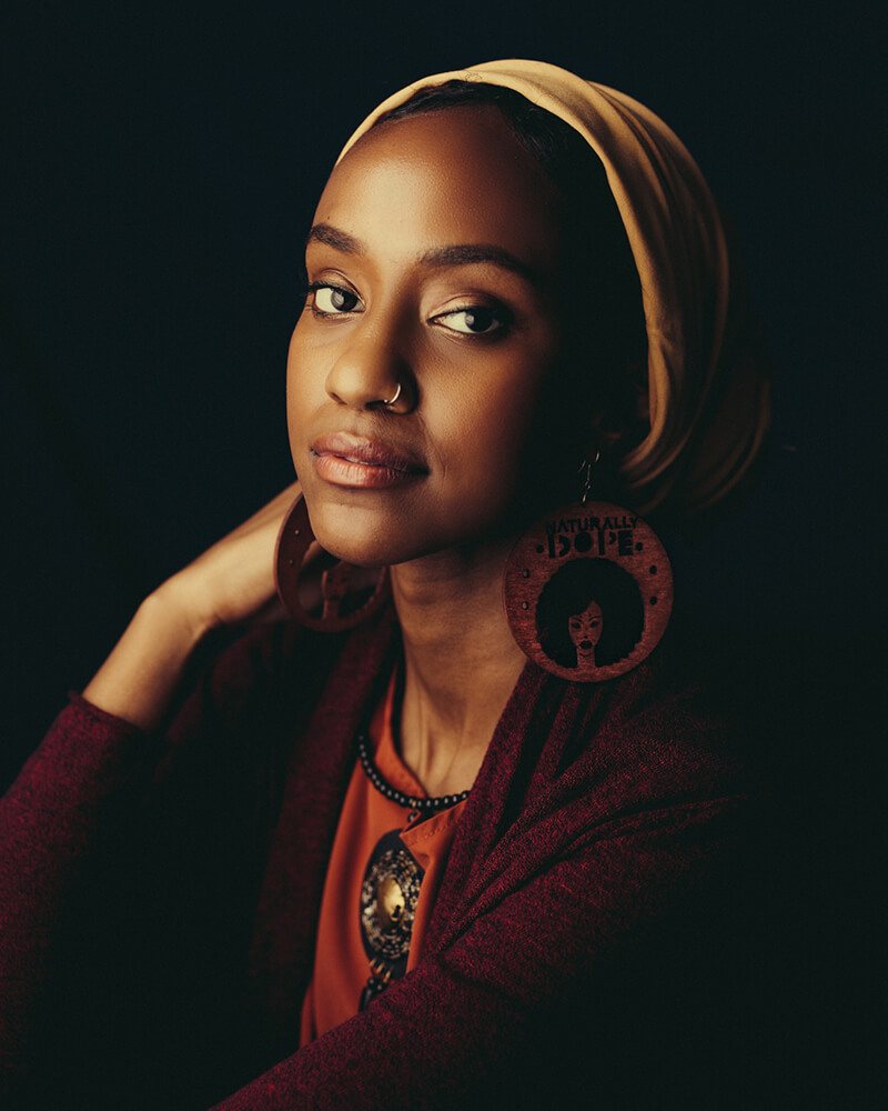 A Black femme person dressed in warm hues and a yellow hijab sits against a dark and shadowy backdrop. She looks over her shoulder at the camera with a slight smile.