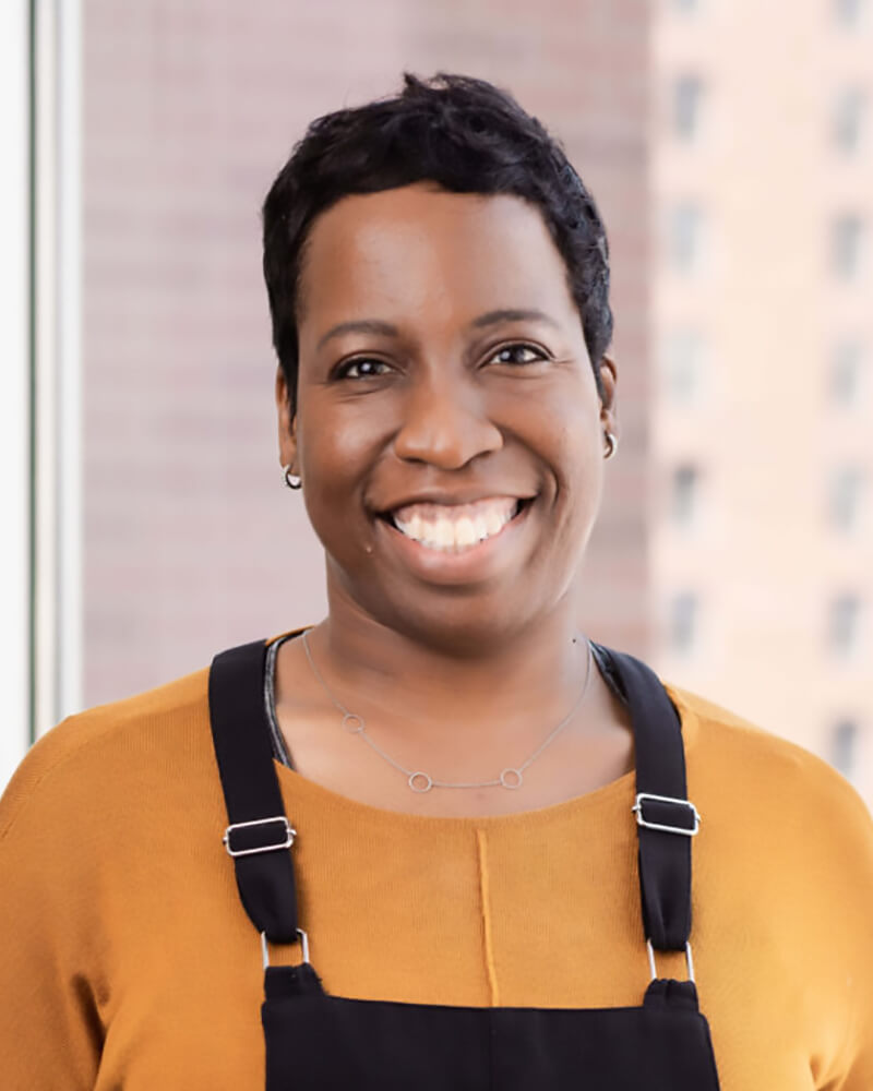 A black disabled woman with short hair, wearing an orange shirt and black overalls is smiling.