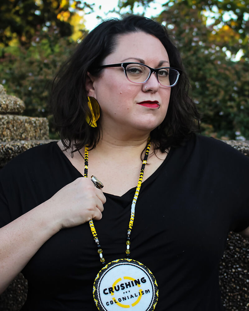 Photo of Jen, a white coded Native, Two-Spirit femme presenting person, with shoulder length brown wavy hair. Jen is wearing a black short-sleeved shirt, black glasses, and an Indigenous made bangle bracelet and beaded medallion that reads 'Crushing Colonialism.' Jen's hands are holding both sides of the medallion and has a tenacious expression on hir face.