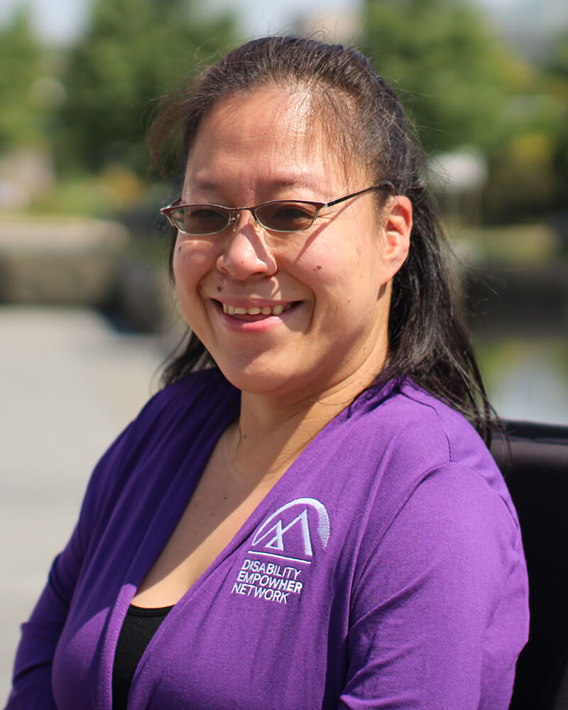 Headshot of a Korean American woman in a power wheelchair smiling, with her shoulder length hair pulled back wearing glasses and a purple shirt with 'Disability EmpowHer Network' logo, seated outside on a sunny day.