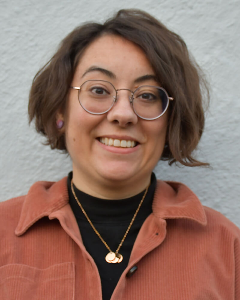 A white person with short brown, wavy hair and round glasses stands against a light blue background. They are smiling and wearing a rose gold necklace with a light brown (almost orange) corduroy shirt over a black top.