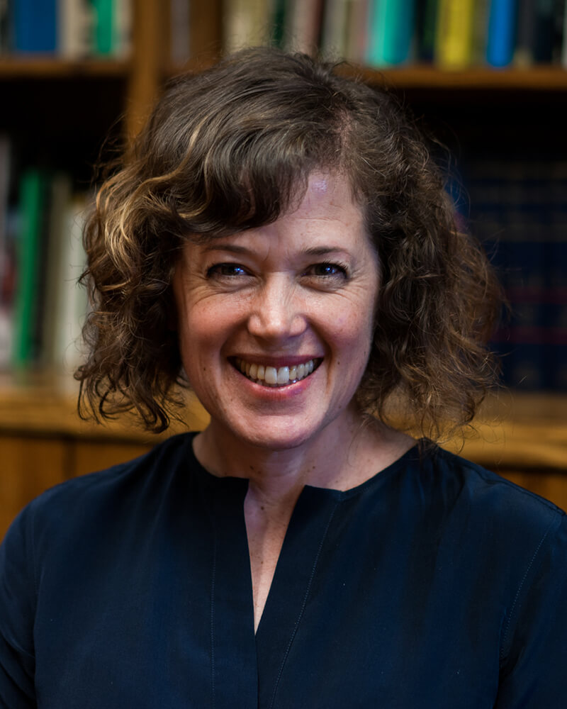 A middle aged white woman with a poufy light-brown bob and a navy blouse sits in front of bookshelves smiling broadly.