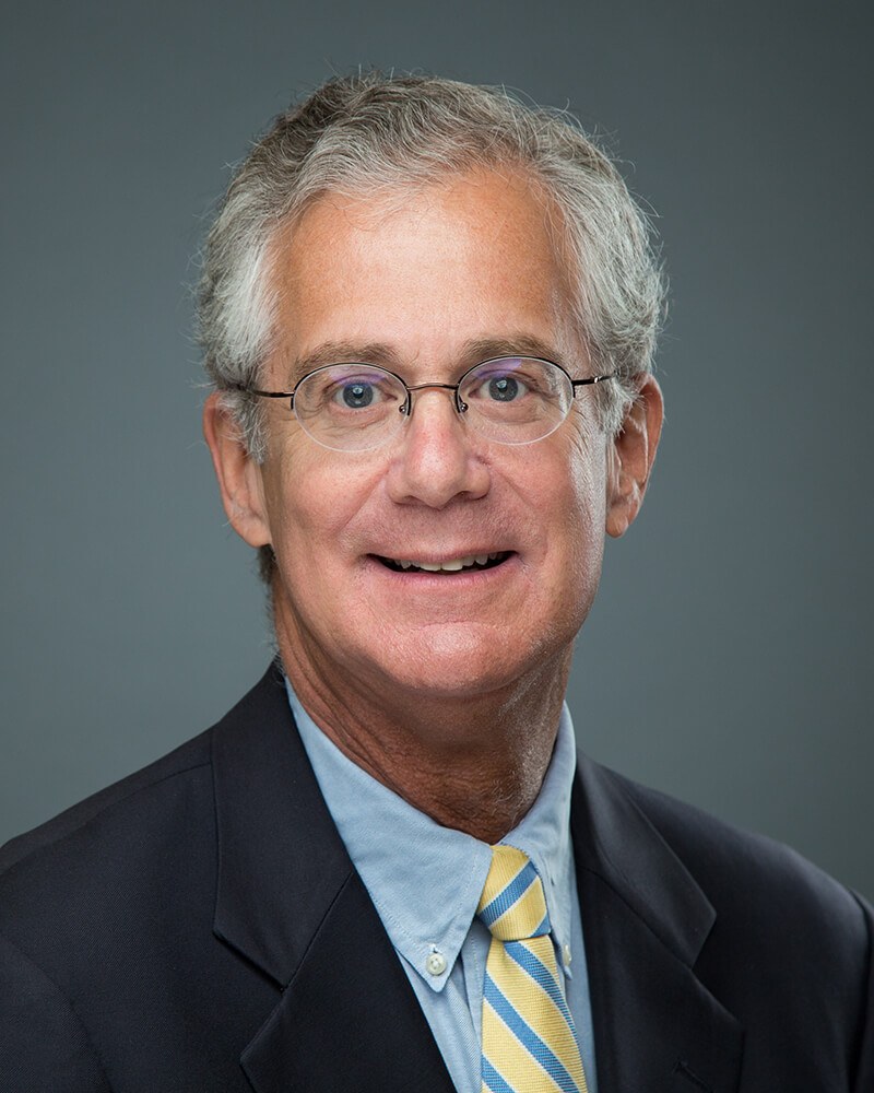 A white man wearing glasses, a light blue shirt, black blazer and yellow & blue stiped tie smiles.