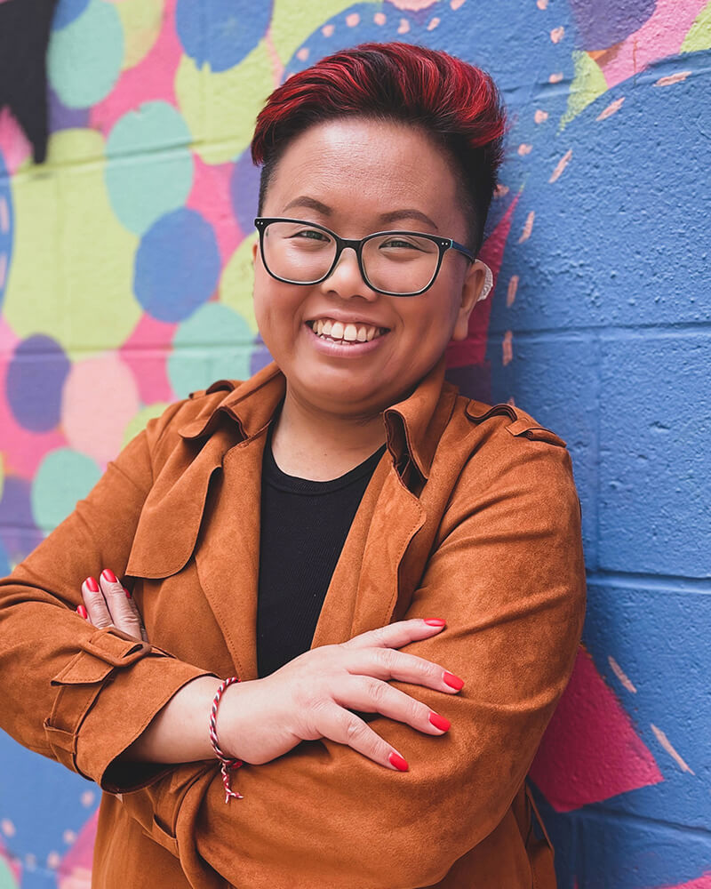 Nancy, a Southeast Asian person, leaning on a brick wall with a colorful mural. Their arms are folded and they are smiling. They have bright red hair and nails and are wearing glasses and an orange jacket.