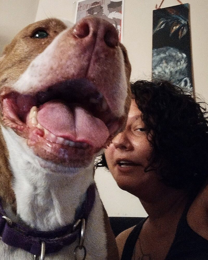 Na - Ha - Ma & their dog Sa-meer-a are sitting on a dark blue couch in their home. Nechama has a round face, olive skin and dark curly hair down to their chin. Samira, a brown and white pitbull with a very big smile and her face, takes up most of the picture frame.