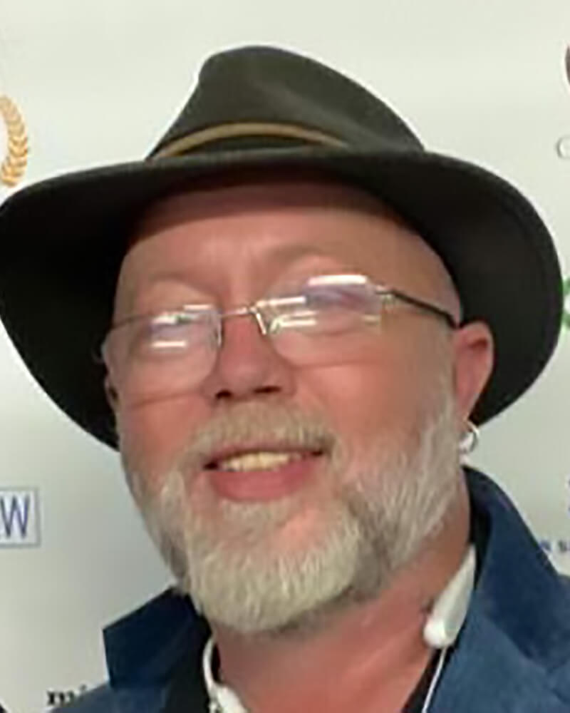 A white man with a beard is wearing glasses and a black hat.